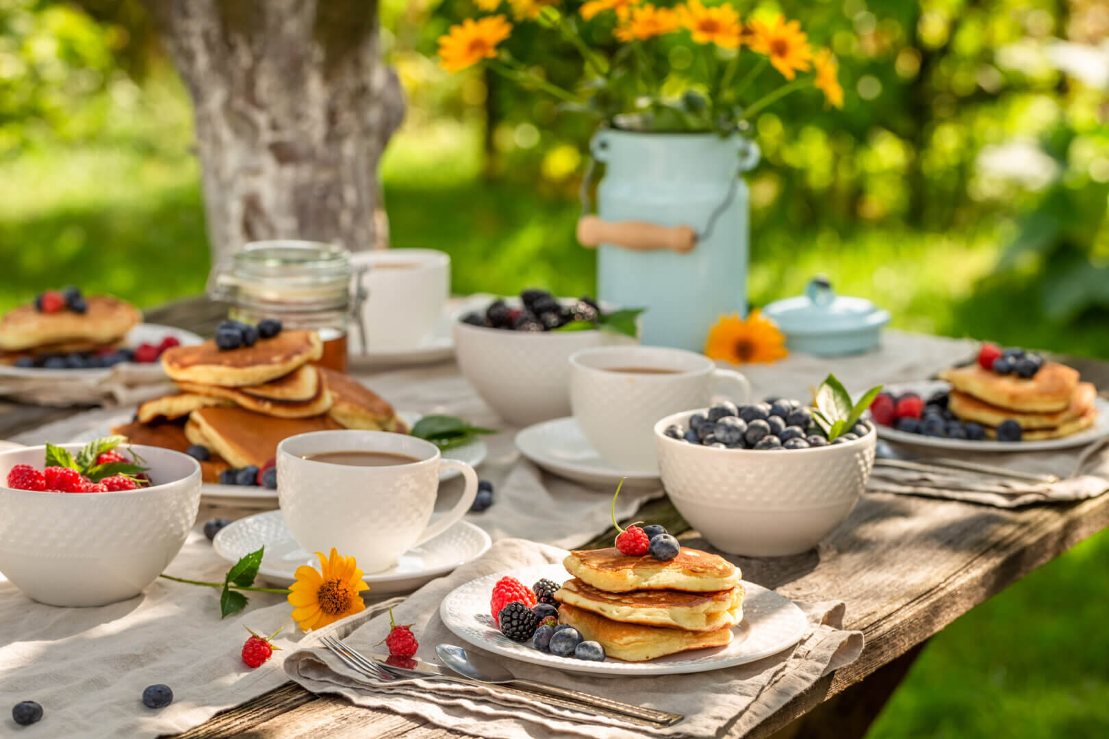 Sommerliches frühstück auf einem outdoortisch aus holz mit pancakes, frischen beeren und kaffee im grünen. Perfekter holztisch für garten und terrasse.