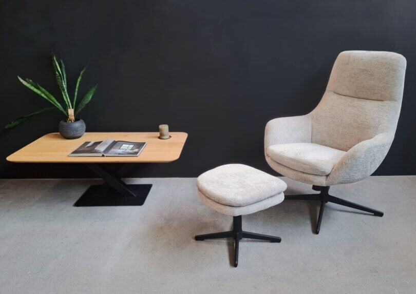 Loungechair mit hocker in heller stofffarbe vor dunkler wand mit couchtisch.