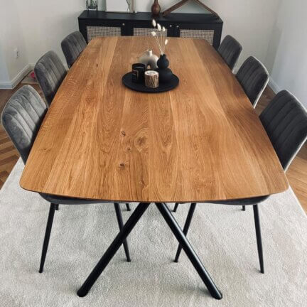 Großer esstisch mit leicht ovaler tischplatte aus eichenholz, aufgestellt auf einem modernen schwarzen gestell mit mehreren stühlen rundherum, platziert auf einem hellen teppich in einem eleganten wohnraum.